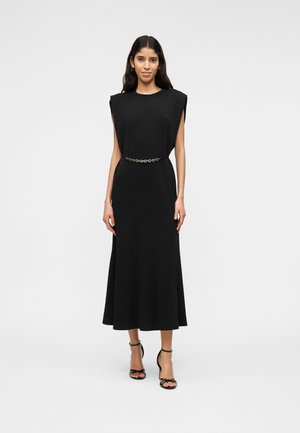 Vrouw draagt een mouwloze zwarte midi-jurk met gewatteerde schouders, een dunne kettingriem en zwarte sandalen met hoge hakken, staand tegen een witte achtergrond.