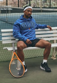 Moletom azul da Nike com grande logotipo, calções pretos e sapatilhas pretas da Nike. Uma raquete de ténis com um cabo laranja repousa ao lado numa banca branca.