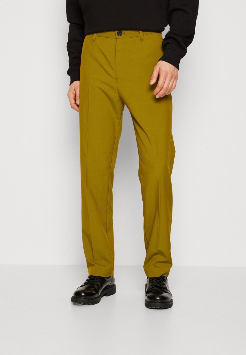 Homme portant un pantalon droit jaune moutarde et des chaussures noires à lacets, debout sur un fond blanc uni.