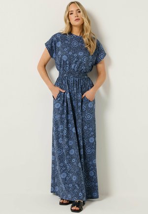 Blonde vrouw staat met handen in de zakken, draagt een blauwe maxi-jurk met bloemenpatroon en zwarte sandalen met open tenen tegen een effen achtergrond.