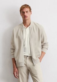 Light beige bomber jacket with ribbed cuffs and collar, worn over a white polo shirt, paired with light beige pants. Smooth texture, zip closure.
