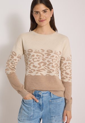 Femme portant un pull beige et brun clair à imprimé léopard avec un jean bleu, debout avec une main dans la poche devant un fond uni.