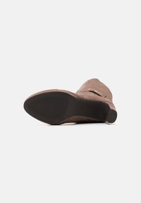 Hnědé semišové kozačky do poloviny stehen s hladkým povrchem, špičaté špičky a plochou gumovou podrážkou. Obsahují malý kovový přezkový detail.