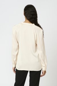 Lys beige langærmet skjorte med struktureret stof, afrundet kant og subtile folder ved skuldrene, parret med sorte bukser.
