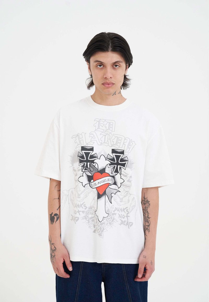 Maglietta in cotone bianco con vestibilità oversize, caratterizzata da grafiche nere a forma di croce e un cuore rosso con la scritta "LOS ANGELES" sul davanti. Design casual.