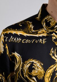 Maglietta nera a maniche corte con un motivo dorato a spirale e testo bianco "JEANS COUTURE", caratterizzata da un colletto classico e una superficie liscia.