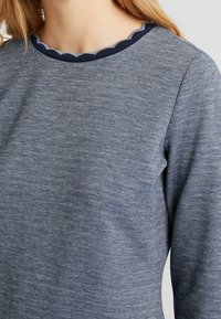 Blå långärmad topp med en texturerad tyg och en scallop-mönstrad marinblå kant i halsringningen. Närbild som framhäver form och detalj.