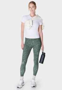 Vrouw in een wit T-shirt, groene, gebloemde leggings en sneakers houdt een zwarte waterfles met de tekst "Sweaty Betty" vast, een crèmekleurige sweater over haar schouders gedrapeerd.