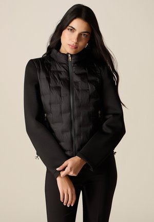 Junge Frau mit langen dunklen Haaren, trägt eine schwarze gesteppte Weste mit Reißverschluss und ein schwarzes langärmeliges Top, steht vor einem beigen Hintergrund.