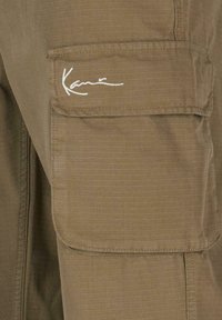 Oliivroheline kangast cargo-püksid, millel on tekstuuriga ruuduline muster, küljetasku ja kontrastne valge logo.