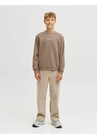 Sudadera marrón con logotipo blanco, pantalones de pana beige y zapatillas grises. El modelo está en un fondo neutro con una pose relajada.