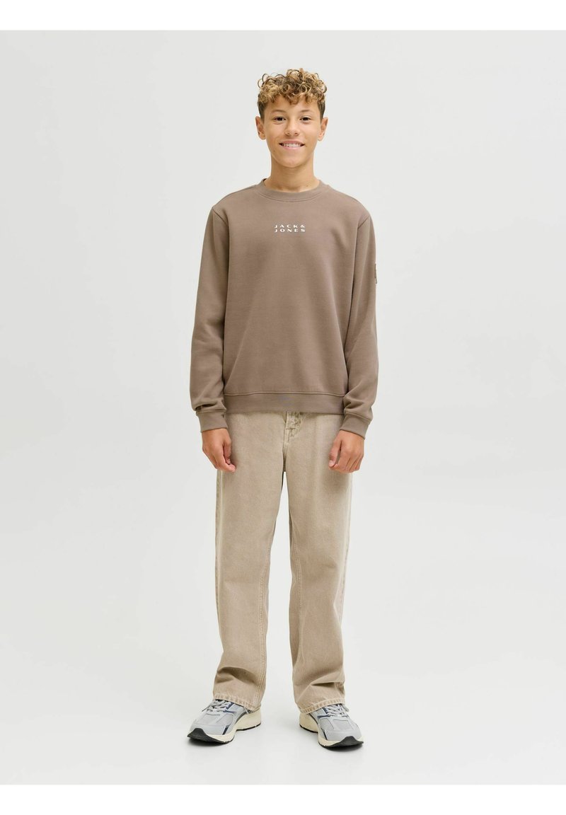 Sudadera marrón con logotipo blanco, pantalones de pana beige y zapatillas grises. El modelo está en un fondo neutro con una pose relajada.
