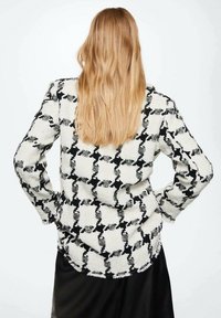 Blazer a rayas en blanco y negro con una tela texturizada, bordes deshilachados y un ajuste entallado, visto desde atrás. Se visible cabello largo y rubio.