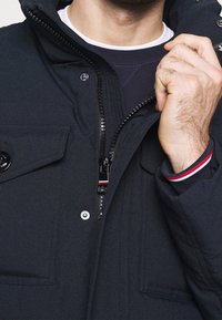 Mörkblå jacka med hög krage, med en framdragbar blixtlås, knäppta fickor och röd-vita randiga detaljer på ärmsluten.