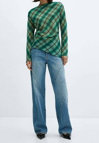 Donna che indossa una maglietta lunga a maniche lunghe asimmetrica a quadri verdi, jeans a gamba larga blu con dettagli delle cuciture e scarpe nere a punta.