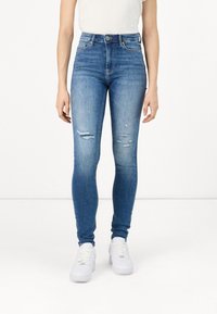 Tievās zils džinsos, kas izgatavoti no denim, ar augstu jostasvietu, viegli nolietotiem elementiem un izplēstiem akcentiem. Pāris ar baltām sporta kedām.