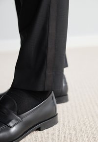 Pantaloni neri su misura presentano un sottile motivo a quadri e una striscia laterale nera a contrasto, abbinati a mocassini neri lucidi e calzini neri.