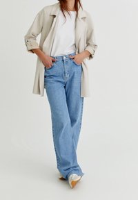 Lysbeige trenchcoat over en hvid t-shirt, parret med højtaljede blå denimjeans med en afslappet pasform og subtile slidte kanter.