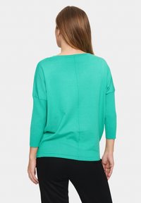 Žalias megztas viršus, laisvas kirpimas, tris ketvirčius ilgio rankovės ir apvali iškirptė. Galinėje pusėje yra vertikalus siūlas.