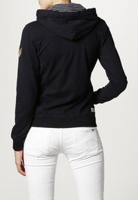Sudadera azul marino con forro interior a rayas, puños y dobladillo acanalados, con un emblema dorado en el hombro izquierdo, llevada sobre pantalones blancos.