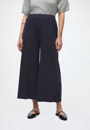 Personne portant un pantalon large noir, une chemise grise à manches courtes, des chaussures plates noires, debout devant un fond clair uni.