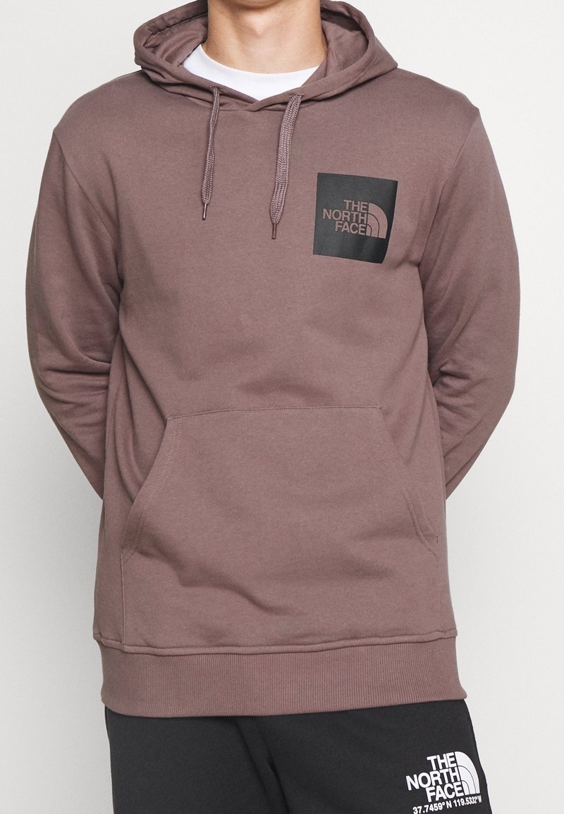Sudadera con capucha marrón hecha de tela suave, con un bolsillo frontal y un parche cuadrado de logo en el área del pecho izquierdo.