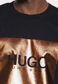 Schwarzes T-Shirt mit einem metallisch bronzefarbenen Rechteck, auf dem der Text "HUGO" und "HUGO BOSS" in fetten schwarzen Buchstaben steht. Kurze Ärmel, runder Ausschnitt.