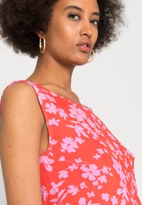 Top sem mangas vermelho com flores grandes em rosa claro, gola redonda e uma textura suave e macia. Brincos em argola dourados complementam o visual.