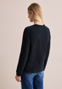 Marineblauer Strickpullover mit strukturierten, abstrakten Mustern. Rundhalsausschnitt und lange Ärmel. Kombiniert mit hellblauen Jeans. Rückansicht dargestellt.