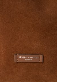 Bruin suède oppervlak met een gladde textuur. Bevat een leren label dat is gecodeerd met "GIANNI CHIARINI FIRENZE" in zilveren letters.