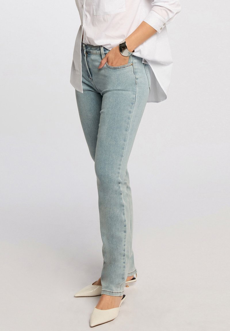 Vaqueros de mezclilla azul claro con un ajuste entallado, textura ligeramente descolorida, combinados con una camisa blanca de botones y zuecos blancos de punta afilada.