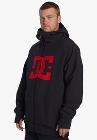 Čierna zipová bunda s veľkým červeným logom DC na prednej strane, s vysokým golierom, nastaviteľnou kapucňou a rebrovanými manžetami a lemom.