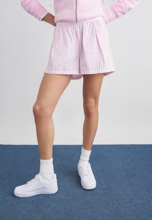 Osoba ubrana w różowo-białe pasiasty szorty, jasnoróżową bluzę z kapturem i zamkiem, białe skarpety do kostek oraz białe trampki stojąca na niebieskim dywanie.