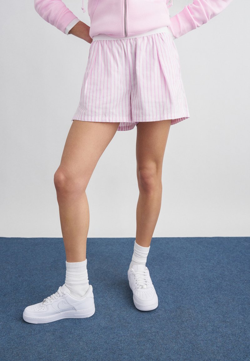 Άτομο που φοράει ροζ και λευκό ριγέ σορτς, ανοιχτό ροζ φούτερ με φερμουάρ, λευκές κάλτσες και λευκά αθλητικά παπούτσια, στέκεται σε μπλε χαλί.