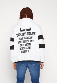 Felpa bianca con testo nero che riporta "TOMMY JEANS" e nomi di città. Strisce nere sulle maniche e vita a coste. Materiale in morbido cotone.