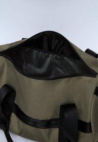 Sac de sport vert olive avec des accents noirs, ouverture zippée et doublure intérieure texturée. Doté de poignées en toile noires robustes.