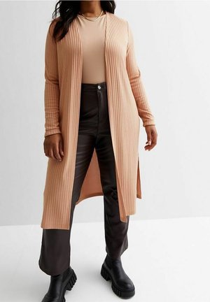Kobieta w długim brzoskwiniowym prążkowanym kardiganie, na beżowej bluzce, czarnych spodniach z wysokim stanem oraz czarnych masywnych botkach na platformie, stojąca na tle białej ściany.
