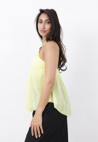 Mujer con cabello largo y oscuro que lleva un top halter sin mangas de color amarillo pálido y pantalones negros, de pie de lado contra un fondo blanco.