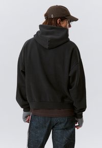 Schwarzer übergroßer Hoodie aus Baumwolle, mit einer großen Kapuze und geripptem Saum. Getragen über einem braunen Shirt und strukturierten blauen Jeans.