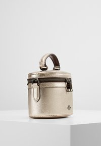 Okrągła, metaliczna złota torba o teksturowanym wykończeniu, z brązowym uchwytem, zamknięciem na zamek i eleganckim designem. Udekorowana czarnymi akcentami.