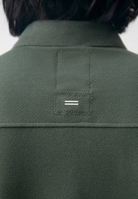Personne portant une veste texturée vert foncé avec un patch rectangulaire cousu et deux bandes blanches horizontales sur le haut du dos.
