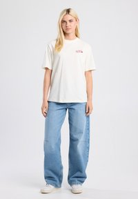 Cream short-sleeve t-shirt with red text graphics, paired with light blue wide-leg jeans and white sneakers, displayed in frontal view.