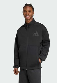 Chaqueta deportiva negra con un cuello alto, cremallera frontal, grandes bolsillos y logotipos en tono. Fabricada con tejido suave y con un corte relajado.