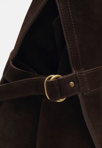 Sac à main en daim marron avec une boucle dorée, des détails cousus et une texture douce. L'intérieur est sombre et spacieux.