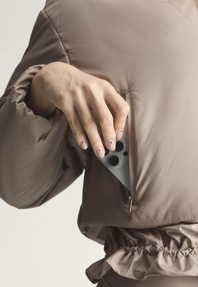 Hand mit minimaler Nagelkunst legt ein Smartphone mit Dreifachkamera in die Reißverschlusstasche einer taupefarbenen Steppjacke.