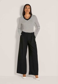 Frau in schwarzen weit geschnittenen Hosen, einem weißen langen gestreiften Oberteil mit Spitzen Ausschnitt und weißen High Heels, vor einem hellen Hintergrund stehend.