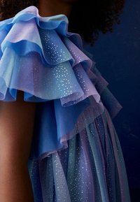 Vaaleansininen ja laventelinvärinen kerroksittain laskostettu helma hopeisilla kimaltavilla yksityiskohdilla. Läpinäkyvä kangas pehmeällä tekstuurilla ja laskeutuva malli.