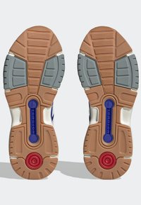 Podešev tenisky s hnědou gumovou základnou, šedými akcenty a červeným kruhovým detailem; vzorované drážky zvyšují trakci a flexibilitu.