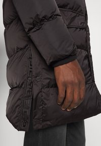 Personne portant un manteau matelassé noir avec une fente latérale à boutons-pression et des poignets côtelés, main détendue à côté d'un pantalon gris foncé.