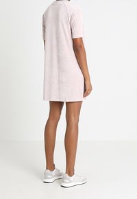 Różowa sukienka o krótkim rękawie, luźnego kroju i miękkiej teksturze, zestawiona z białymi butami sportowymi. Sukienka ma gładki, minimalistyczny design.